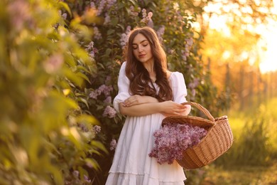 Photo of Beautiful woman with basket of lilac flowers outdoors in morning