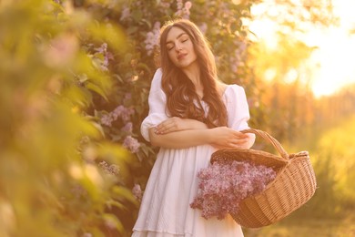 Photo of Beautiful woman with basket of lilac flowers outdoors in morning