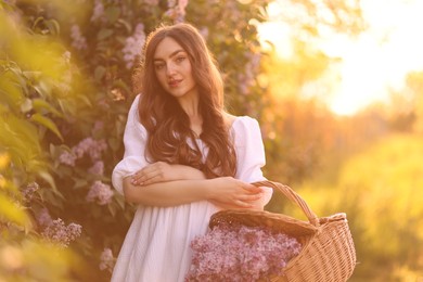 Photo of Beautiful woman with basket of lilac flowers outdoors in morning