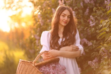 Photo of Beautiful woman with basket of lilac flowers outdoors in morning