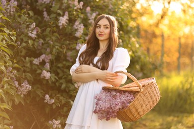Photo of Beautiful woman with basket of lilac flowers outdoors in morning