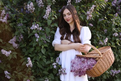 Photo of Beautiful woman with basket of lilac flowers near bush outdoors