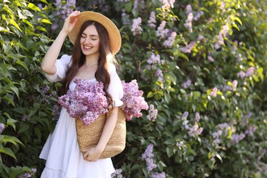 Photo of Smiling woman with bag of lilac flowers near bush outdoors