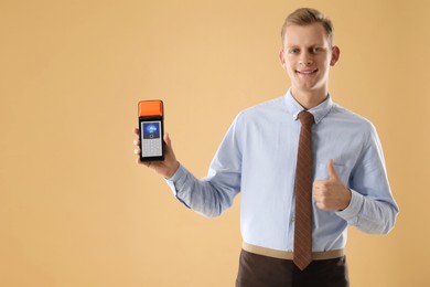 Image of Man holding payment terminal with NFC system and showing thumbs-up on pale brown background