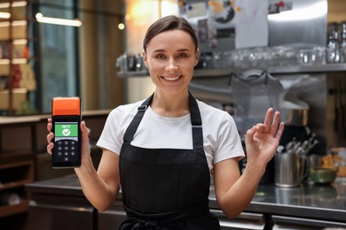 Image of Woman holding payment terminal with NFC system and showing ok in restaurant