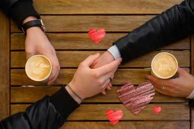 Image of Lovely couple holding hands while drinking coffee at wooden table, top view. Drawn hearts near them