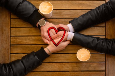 Image of Lovely couple holding hands while drinking coffee at wooden table, top view. Drawn heart near them