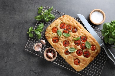 Photo of Delicious focaccia with tomatoes, spices and knife on gray textured table, flat lay