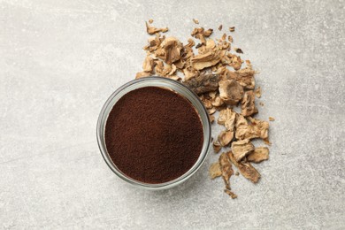 Photo of Chicory powder in bowl and pieces of dry roots on gray textured table, flat lay