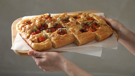 Photo of Woman holding cut delicious focaccia with tomatoes and spices on gray background, closeup