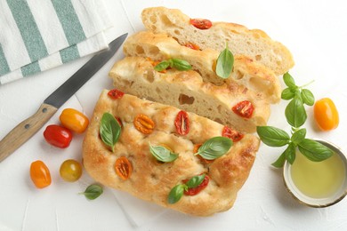 Photo of Cut delicious focaccia with tomatoes, basil, oil and knife on light table, flat lay