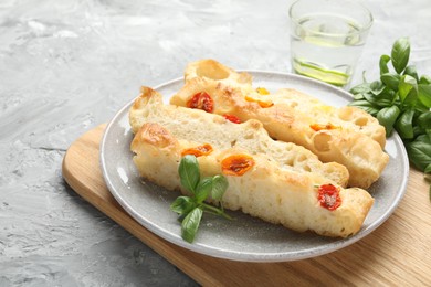 Photo of Pieces of delicious focaccia with tomatoes, basil and glass of water on gray textured table, closeup