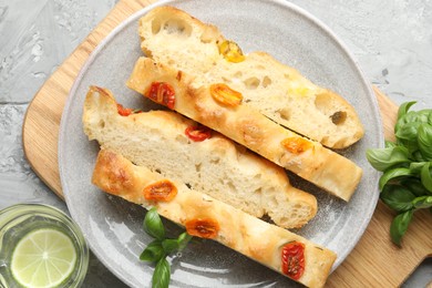 Photo of Pieces of delicious focaccia with tomatoes, basil and glass of water on gray textured table, flat lay