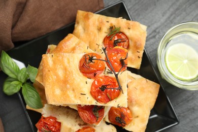 Photo of Pieces of delicious focaccia with tomatoes, spices and glass of water on gray table, flat lay