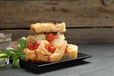 Photo of Pieces of delicious focaccia with tomatoes, basil and glass of water on gray table