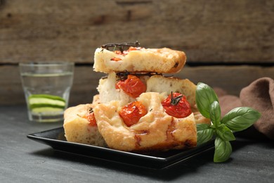 Photo of Pieces of delicious focaccia with tomatoes, basil and glass of water on gray table