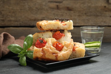 Photo of Pieces of delicious focaccia with tomatoes, basil and glass of water on gray table