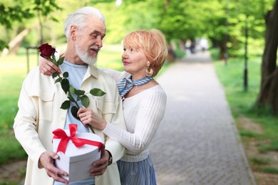 Photo of Lovely senior couple with gift box and rose in park, space for text