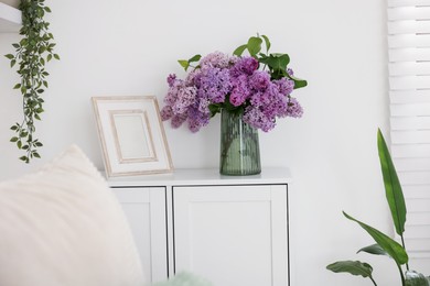 Photo of Beautiful lilac flowers in vase and picture frame on cabinet indoors