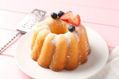 Photo of Tasty Bundt cake with powdered sugar, berries and server on pink wooden table, closeup