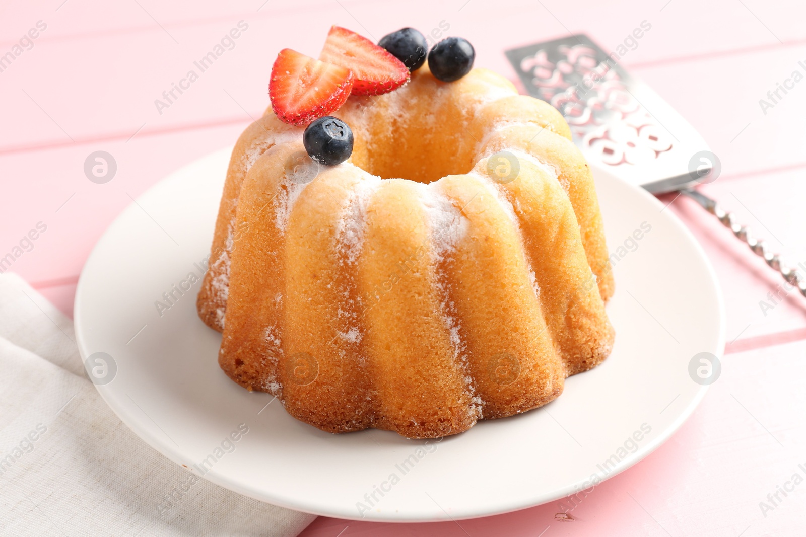 Photo of Tasty Bundt cake with powdered sugar, berries and server on pink wooden table, closeup