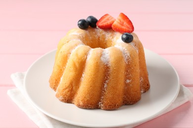 Photo of Tasty Bundt cake with powdered sugar and berries on pink wooden table, closeup