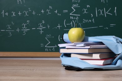 Photo of Backpack, apple and books on wooden table near blackboard with formulas, closeup. Space for text