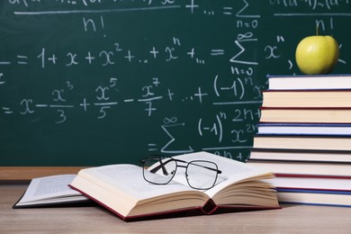 Photo of Books, glasses and apple on wooden table near blackboard with formulas, closeup