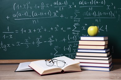 Photo of Books, glasses and apple on wooden table near blackboard with formulas