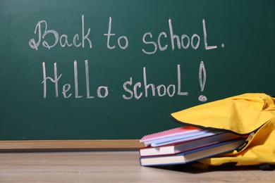 Photo of Phrases Back To School and Hello School with exclamation mark on blackboard. Backpack and books on wooden table