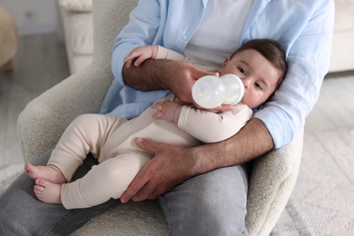 Photo of Father feeding his little baby with bottle on armchair at home, closeup
