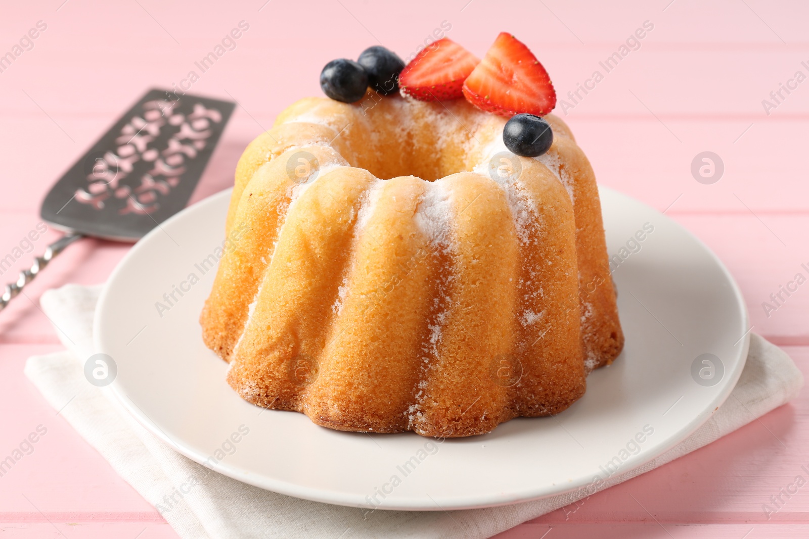 Tasty Bundt cake with powdered sugar, berries and server on pink wooden table, closeup Photo of Tasty Bundt cake with powdered sugar, berries and server on pink wooden table, closeup