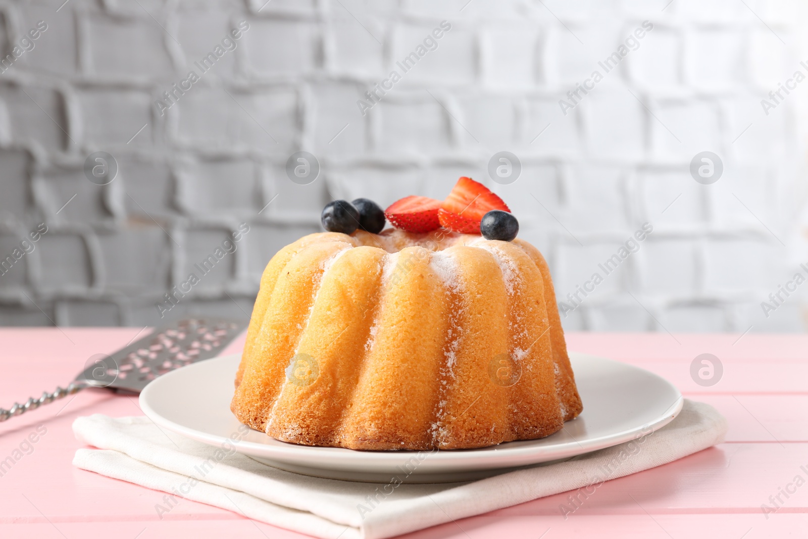 Photo of Tasty Bundt cake with powdered sugar, berries and server on pink wooden table, closeup