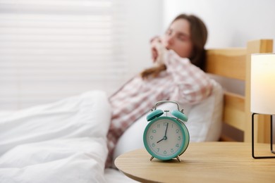 Photo of Young woman yawning on bed indoors, focus on alarm clock