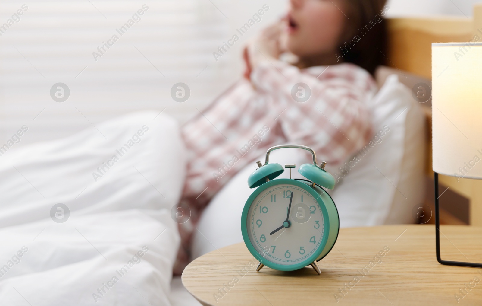 Young woman yawning on bed indoors, focus on alarm clock Photo of Young woman yawning on bed indoors, focus on alarm clock