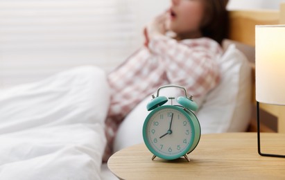Photo of Young woman yawning on bed indoors, focus on alarm clock