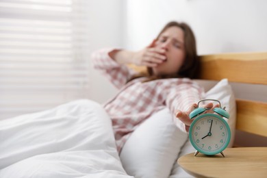 Photo of Young woman yawning and turning off alarm clock on bed at home, selective focus