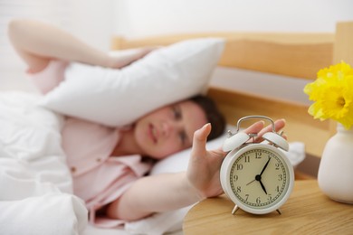 Photo of Frustrated young woman covering ears with pillow and turning off alarm clock on bed at home, selective focus