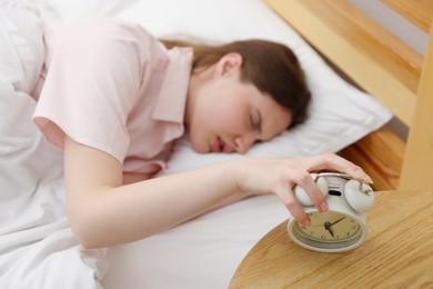 Photo of Frustrated young woman turning off alarm clock on bed at home, selective focus