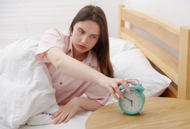 Photo of Frustrated young woman turning off alarm clock on bed at home