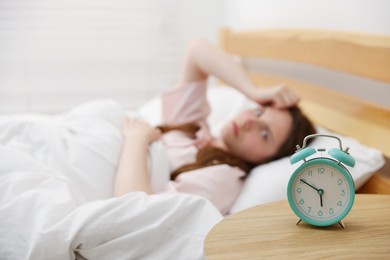 Photo of Frustrated young woman on bed at home, focus on alarm clock