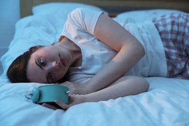 Photo of Frustrated young woman with alarm clock suffering from insomnia on bed indoors
