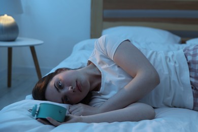 Photo of Frustrated young woman with alarm clock suffering from insomnia on bed indoors