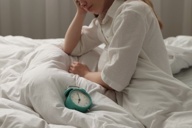 Photo of Upset young woman sitting on bed at home, focus on alarm clock
