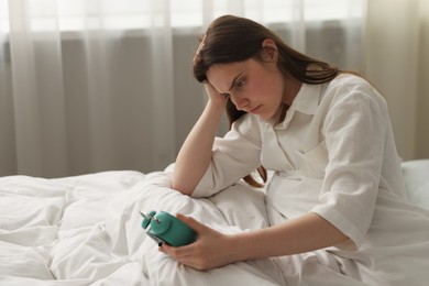 Photo of Upset young woman with alarm clock sitting on bed at home