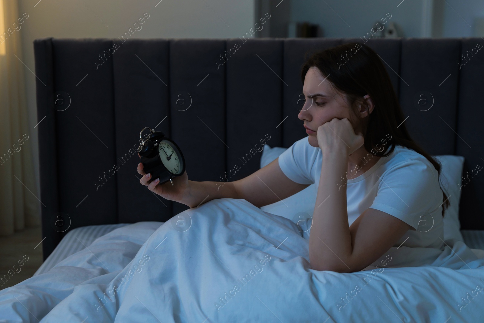 Frustrated young woman with alarm clock suffering from insomnia on bed indoors Photo of Frustrated young woman with alarm clock suffering from insomnia on bed indoors