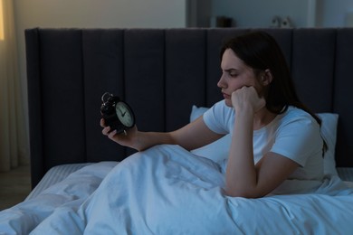 Photo of Frustrated young woman with alarm clock suffering from insomnia on bed indoors