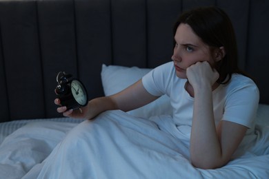 Photo of Frustrated young woman with alarm clock suffering from insomnia on bed indoors