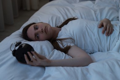 Photo of Frustrated young woman with alarm clock suffering from insomnia on bed indoors