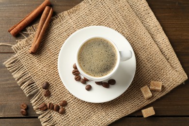 Photo of Aromatic coffee in cup, beans, brown sugar and cinnamon on wooden table, flat lay